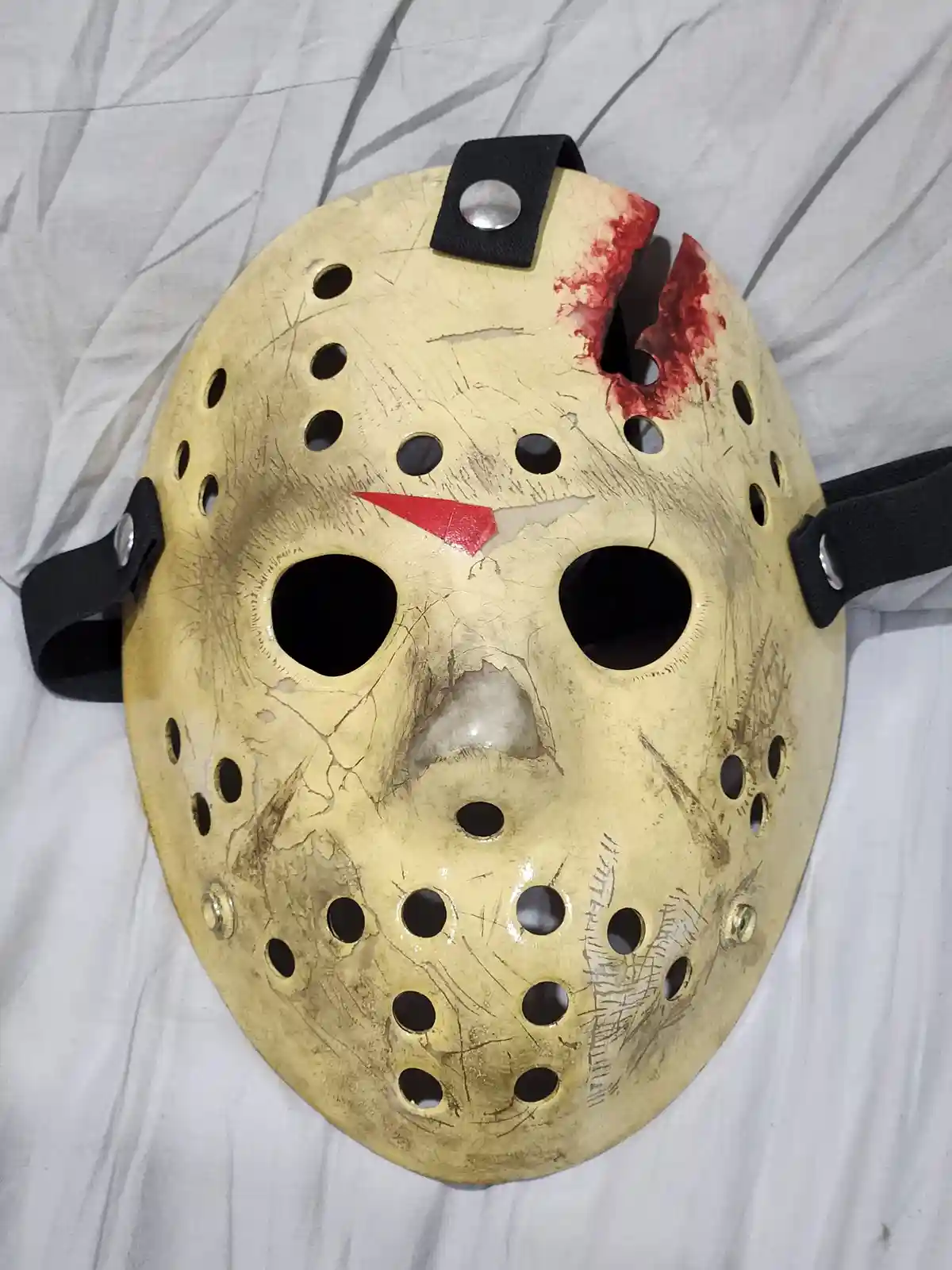A heavily damaged, yellow hockey mask with numerous scratches, a torn right eye area with red paint resembling blood, and a…