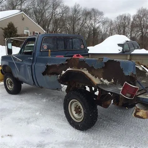 a heavily rusted, blue pickup truck with peeling paint and missing panels sits in a snowy yard, its rear bumper and tailgate…