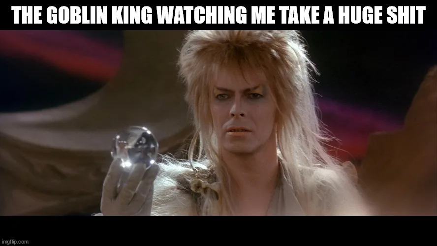 A still image of David Bowie as the Goblin King from Labyrinth, holding a crystal ball with a shocked expression. The…