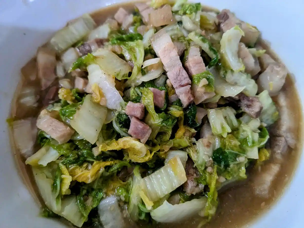 A close-up photo shows a white bowl filled with a dish containing chopped cabbage leaves, pieces of cooked pork, and a…