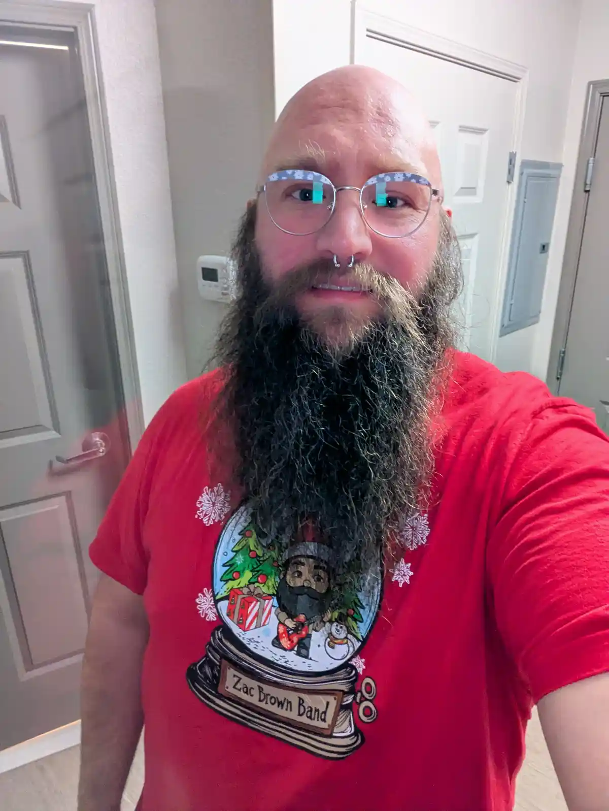 A bald man with a long, dark beard and glasses wearing a red t-shirt featuring a snow globe design with a bearded figure…