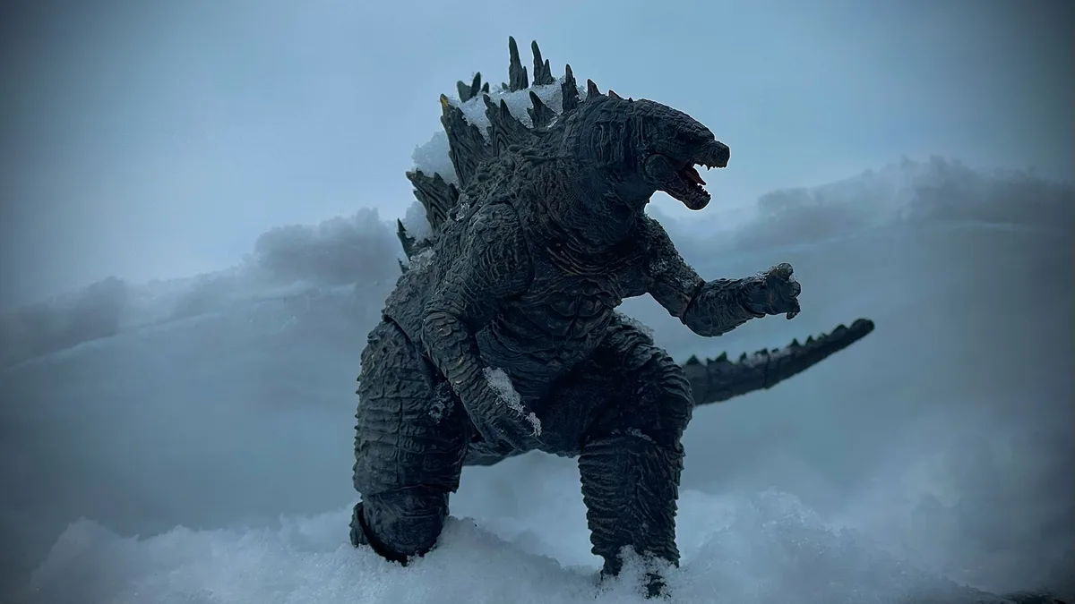 A dark grey Godzilla action figure stands in a snowy, foggy landscape with its mouth open and one arm raised, covered in…