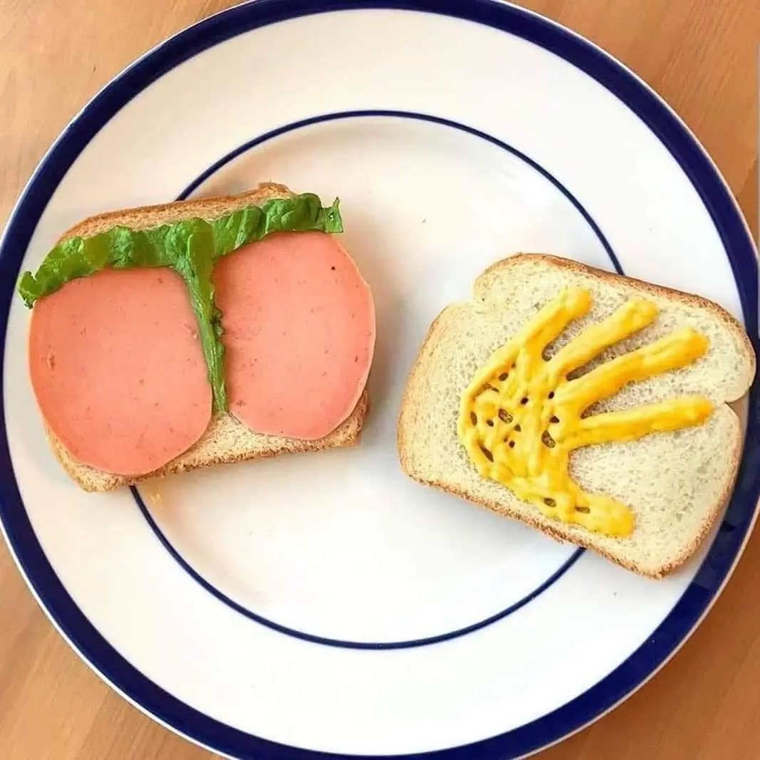 Two slices of bread on a white plate with blue rim sit on a wooden surface. One slice has two round slices of pink deli meat…