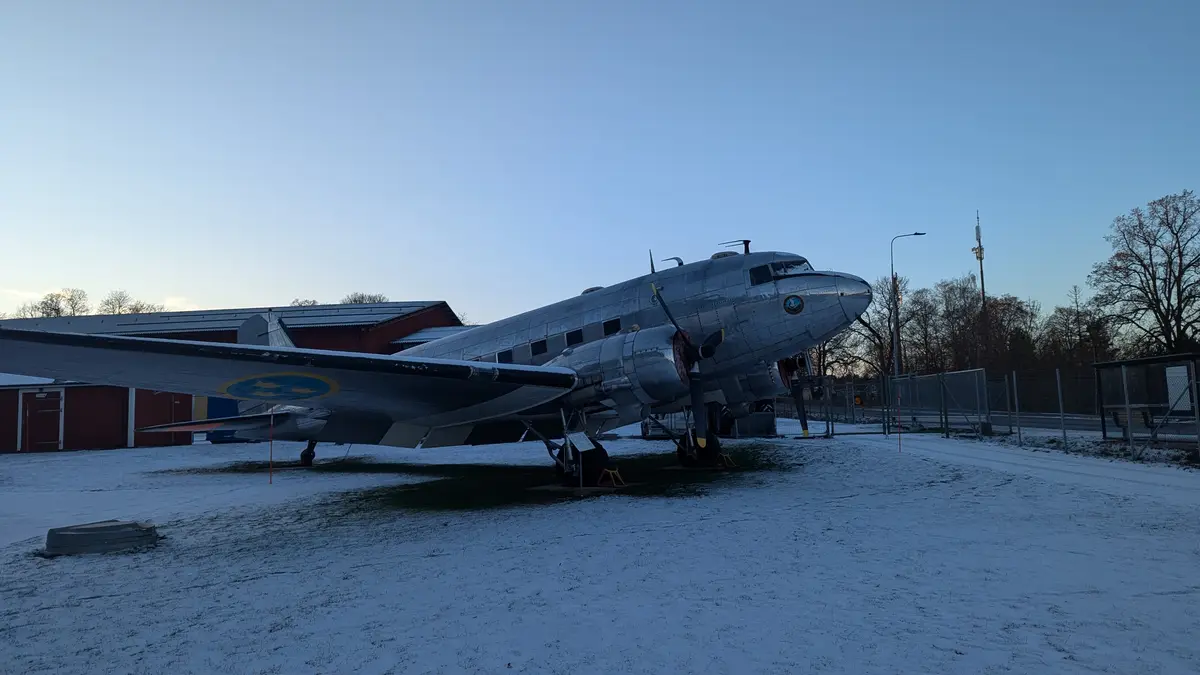 A silver vintage propeller airplane, likely a military transport, is parked outdoors on a snowy field with a red hangar…