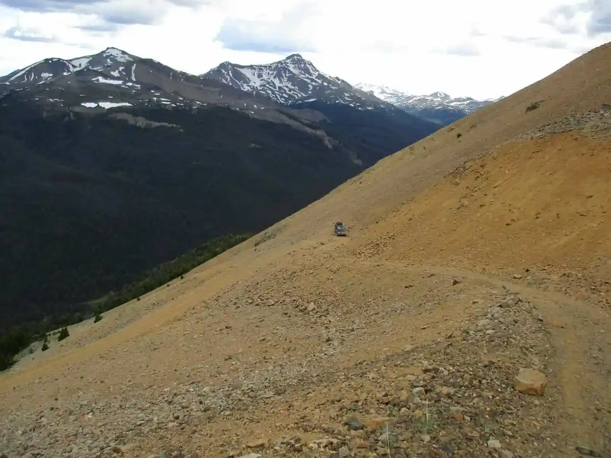 A small vehicle is driving on a steep, rocky dirt road on a mountain slope, with snow-capped peaks and dense forests visible…