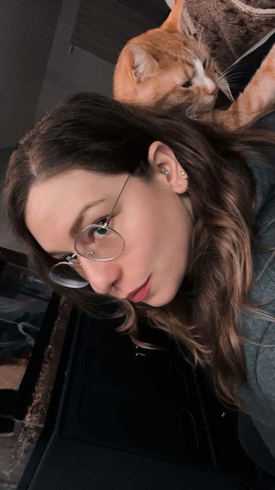 A selfie of a woman with dark hair and round glasses looking at the camera, with an orange tabby cat perched on her head…
