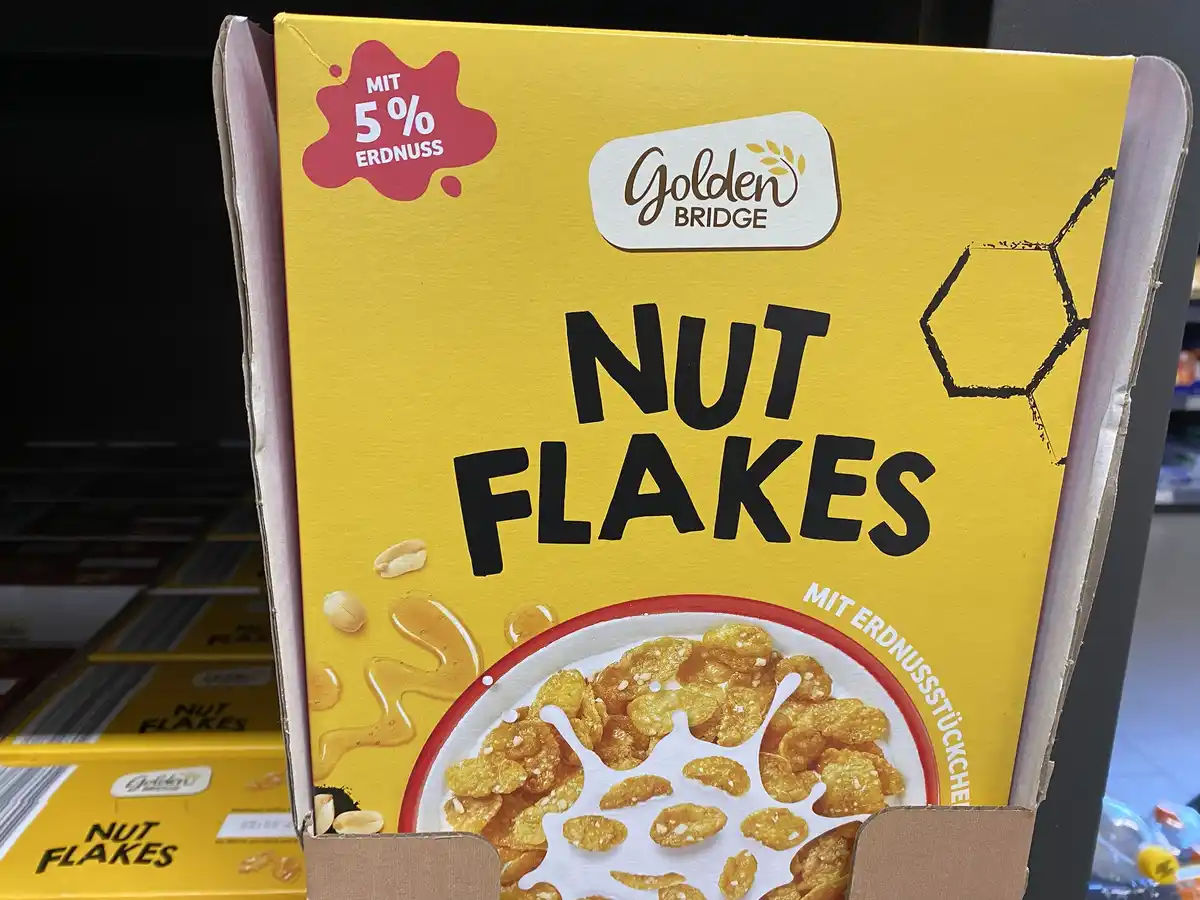 A close-up of a yellow cereal box labeled 'Golden Bridge Nut Flakes' with German text 'mit 5% erdnuss' in a red splash…