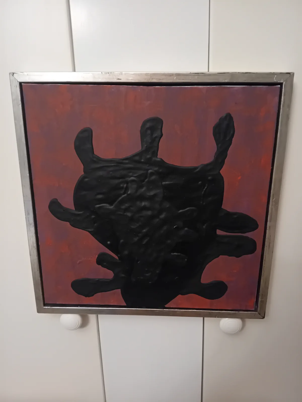 a framed painting with a textured black splat on a red-orange background, mounted on a white wall above two white knobs. the…