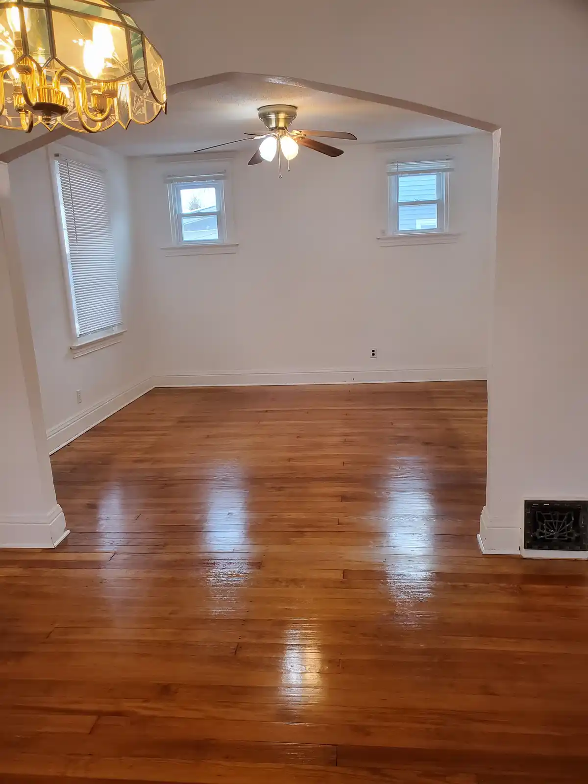 An empty room with hardwood floors, white walls, and three windows, viewed from a doorway. A decorative chandelier hangs in…