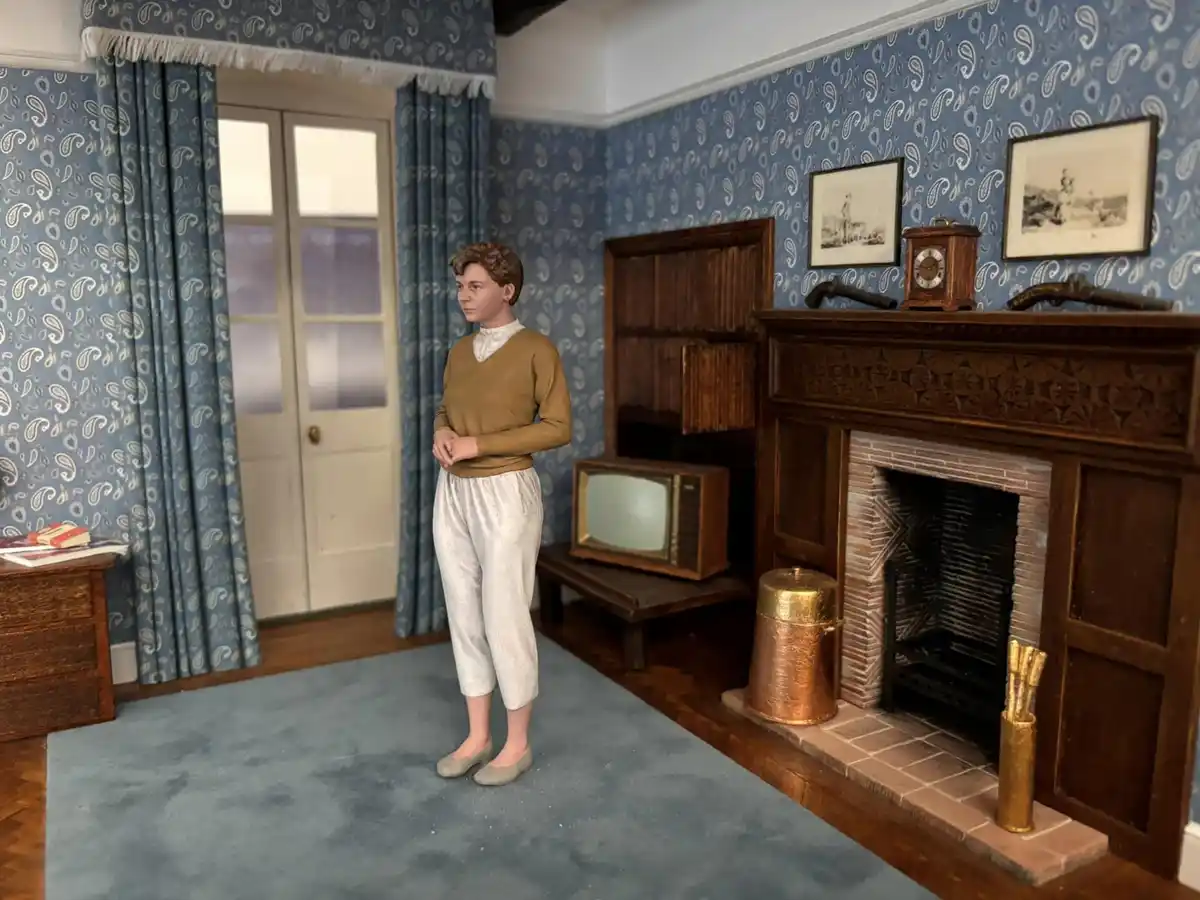 A detailed dollhouse scene shows a doll-like figure standing in a vintage living room with blue paisley wallpaper, a…