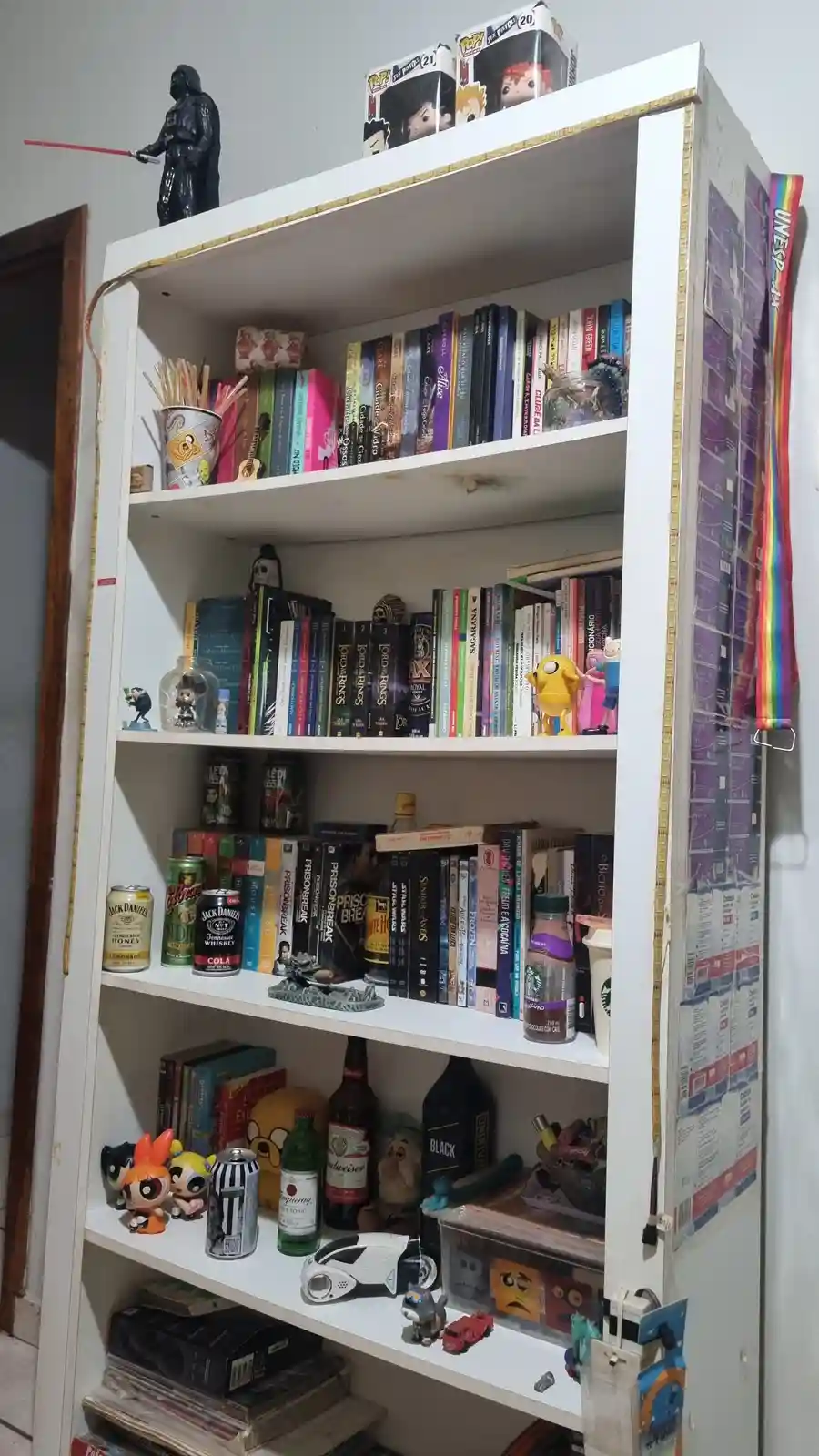 A white bookshelf is filled with books, pop culture figurines like Darth Vader and Powerpuff Girls, drink cans, bottles, and…