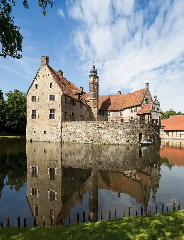 A picturesque stone castle with red-tiled roofs and a central tower stands beside a calm body of water, its reflection…