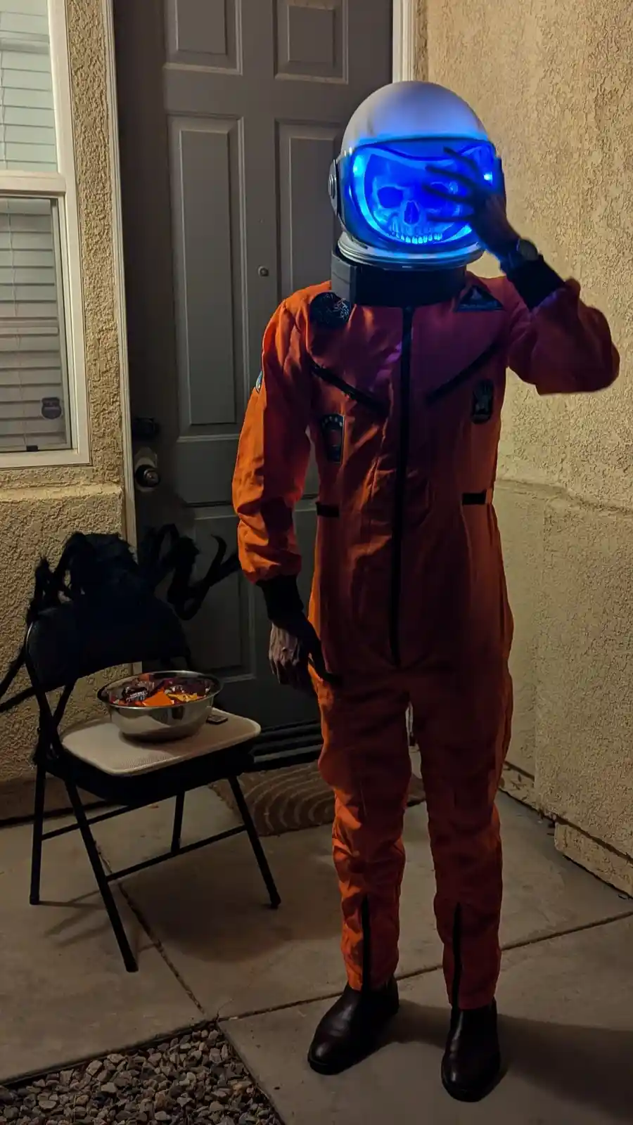 A person in an orange astronaut suit stands outside a house at night, wearing a helmet with a glowing blue skull face…