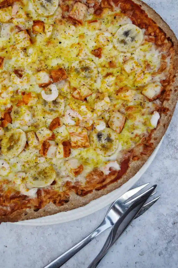 A close-up photo of a pizza with a brown crust, melted cheese, and toppings including sliced bananas, shrimp, and diced…