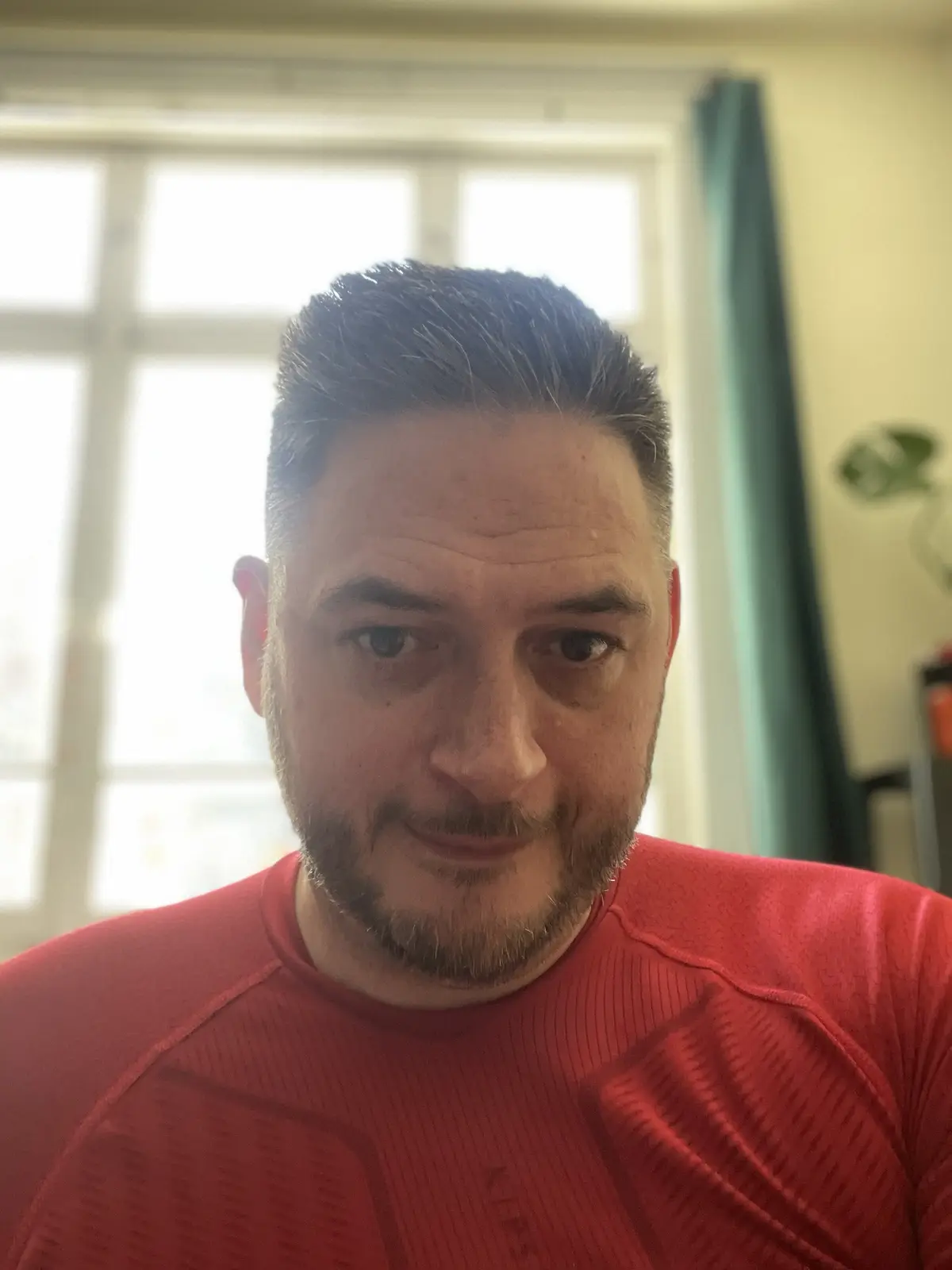 A man with a beard and styled hair, wearing a red athletic shirt, stares directly at the camera with a subtle, smug…