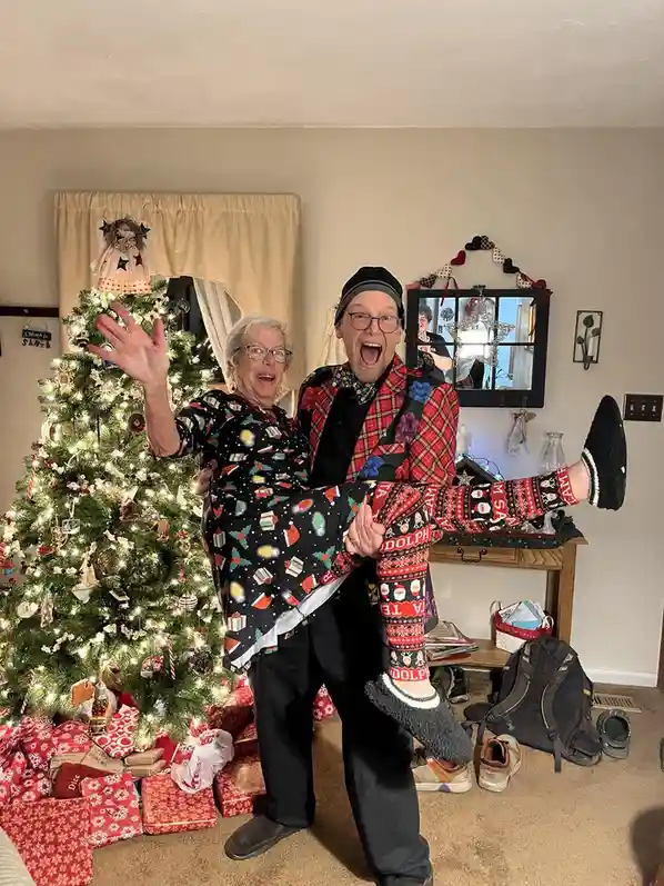 A man in festive red plaid shirt and patterned socks joyfully lifts an older woman in a Christmas-themed pajama set in front…