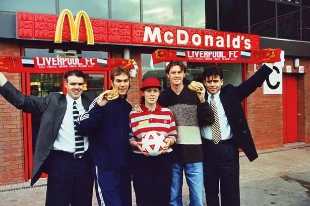 Five men pose in front of a red McDonald's restaurant with Liverpool FC scarves and banners. One man in a red hat holds a…