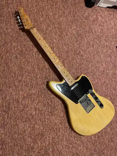 A light-wood electric guitar with a black pickguard lies diagonally on a textured brown carpet. The overlaid text reads 'i…