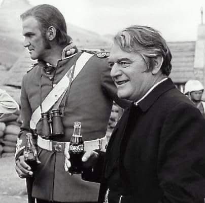 A black-and-white photo shows two men outdoors, one in a military uniform holding binoculars and a bottle, the other in…