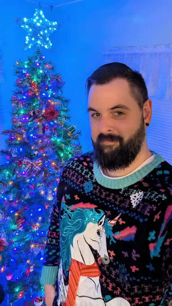 A man with a beard and black ear piercing stands in front of a brightly lit, decorated Christmas tree. He wears a black…