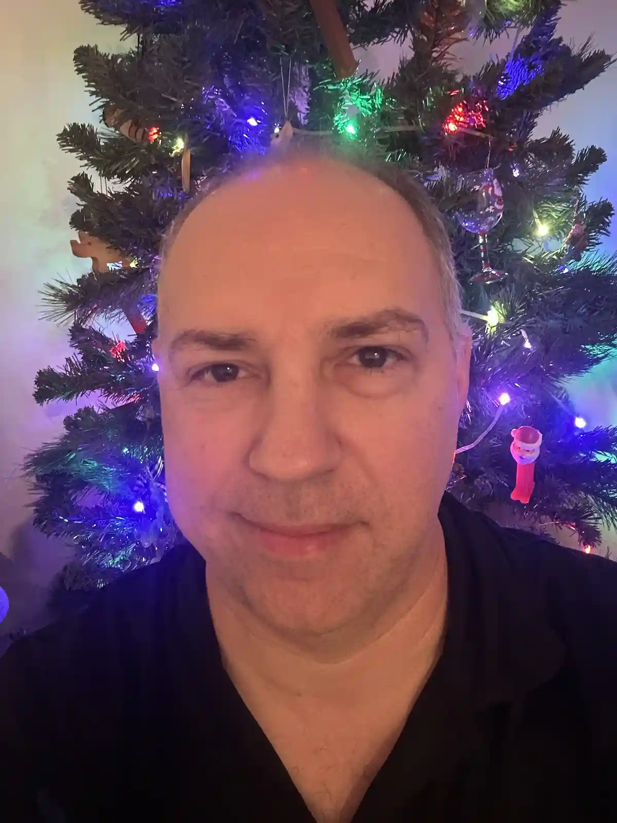 A man with a receding hairline and a slight smile stands in front of a decorated christmas tree with colorful lights and…