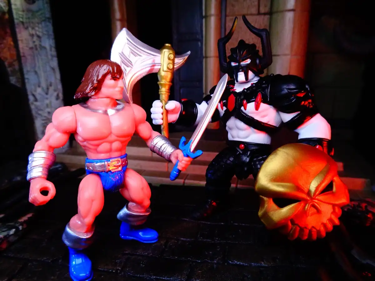 Two action figures face off: a muscular, shirtless hero with blue shorts and a sword, and a dark-armored villain with a…