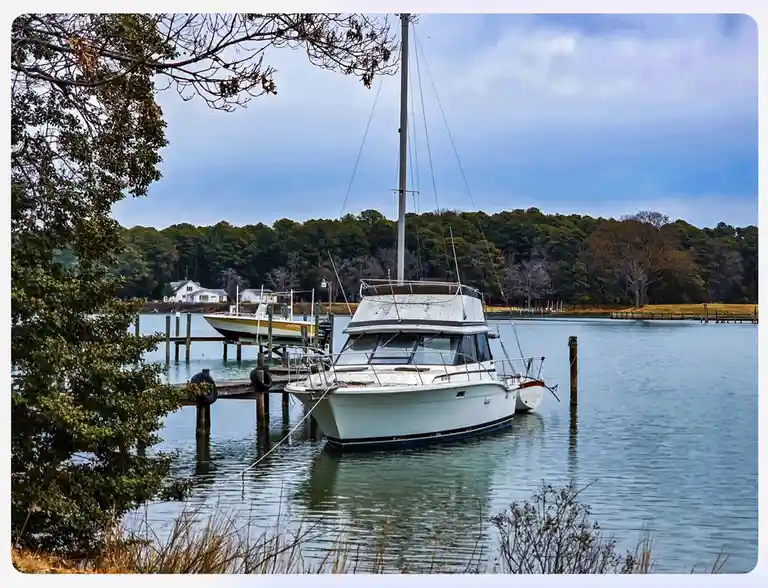 A white sailboat is docked at a wooden pier in calm water, with trees and a house visible in the background under a partly…