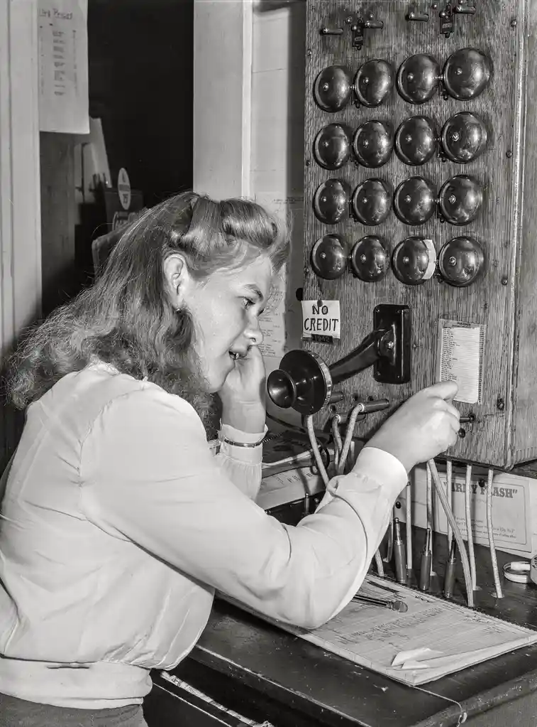 A black-and-white photo from 1942 shows a young woman operating a vintage telephone switchboard, holding the receiver to her…