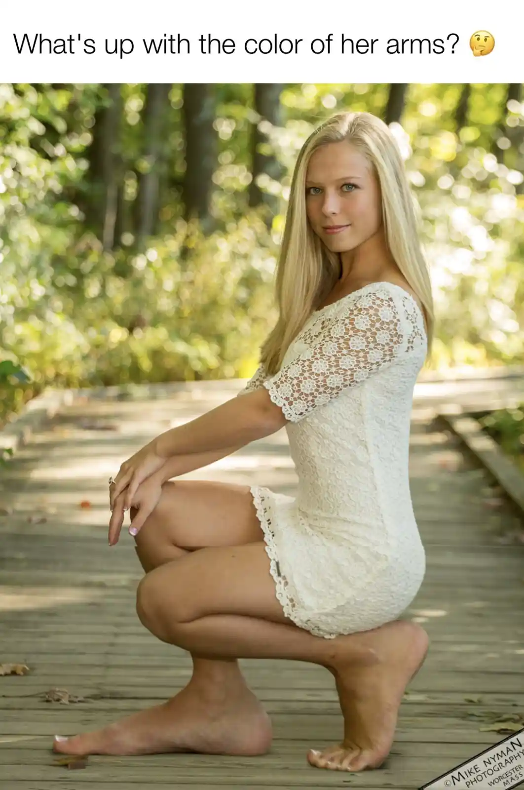 A blonde woman in a white lace dress is squatting barefoot on a wooden boardwalk in a forest. Her arms are visibly darker…