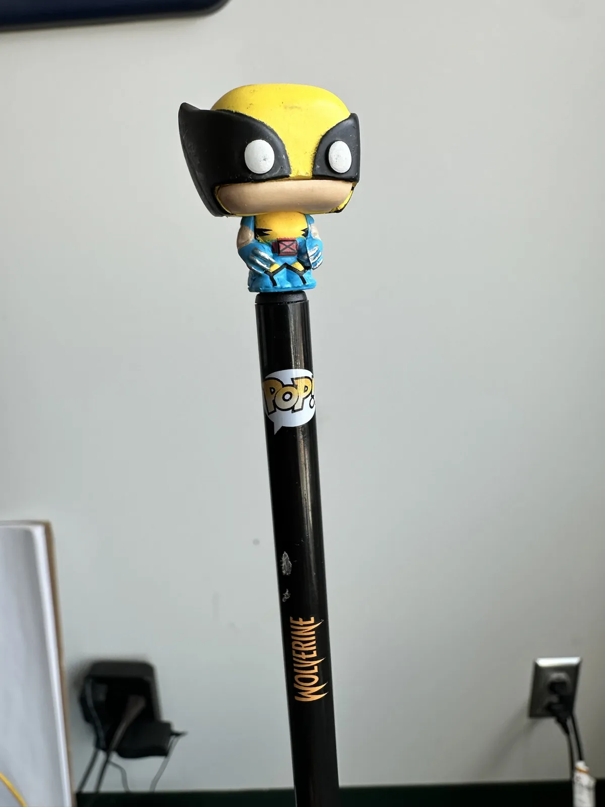 A Funko Pop! figure of Wolverine is perched atop a black pen that has 'WOLVERINE' written vertically in gold lettering. The…