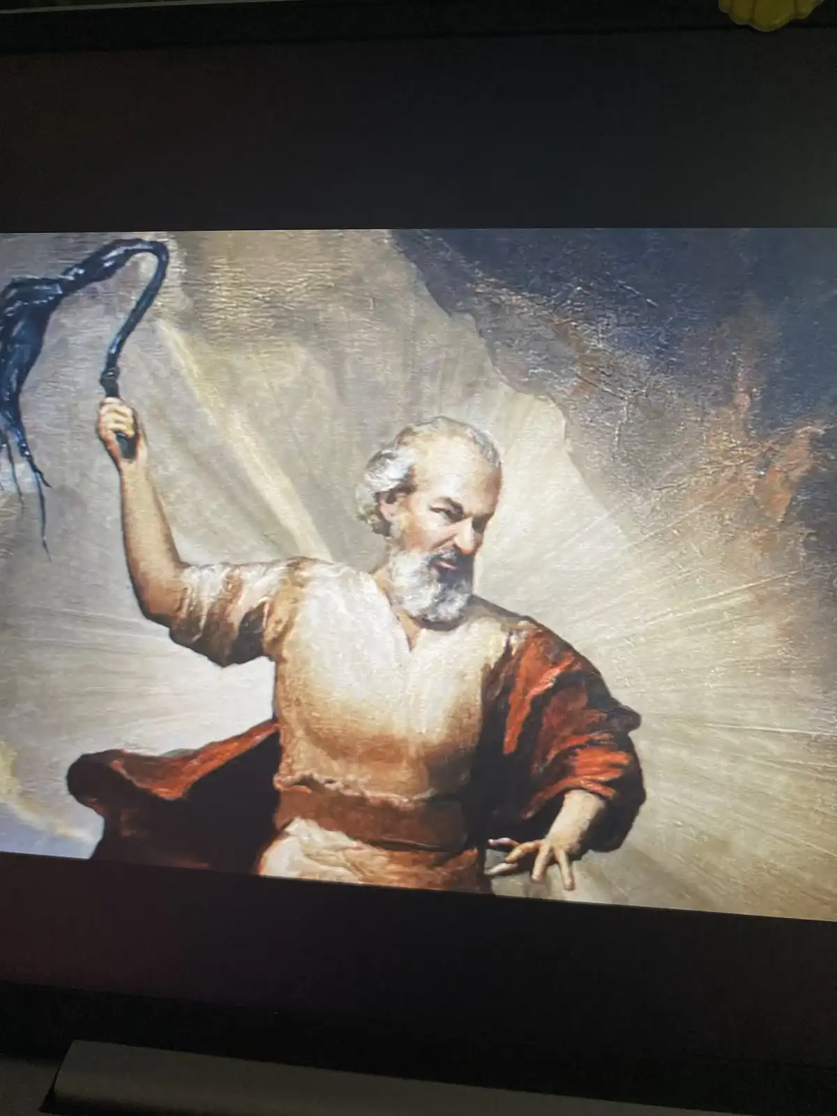 A classical painting depicts a bearded, elderly man in robes holding a black whip aloft, his face contorted in a grimace…
