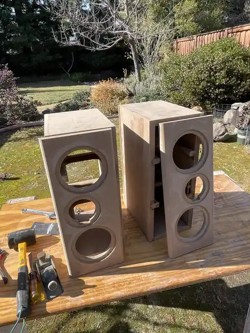 Two unfinished, boxy speaker enclosures made of light brown concrete stand on a wooden workbench outdoors, surrounded by…