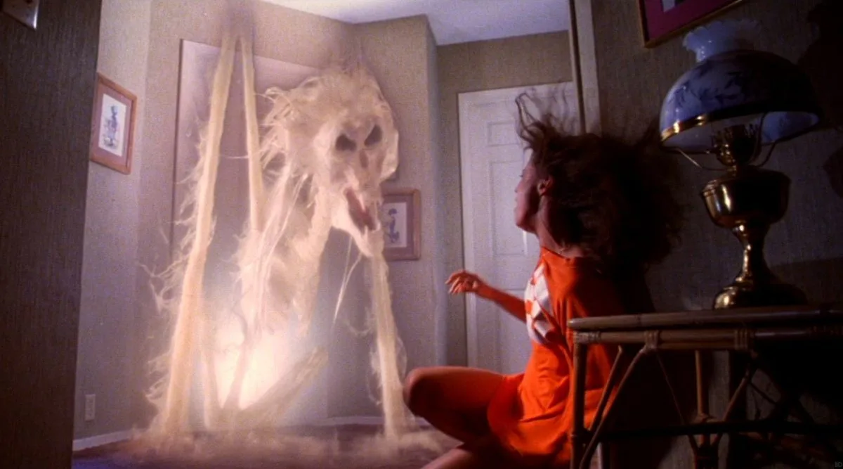 a woman in an orange outfit sits on a chair in a dimly lit room, her hair flying as she stares in terror at a ghostly…
