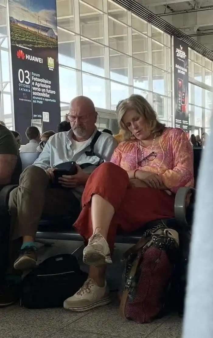 A man and woman are seated side-by-side in what appears to be an airport waiting area, with large windows and advertising…