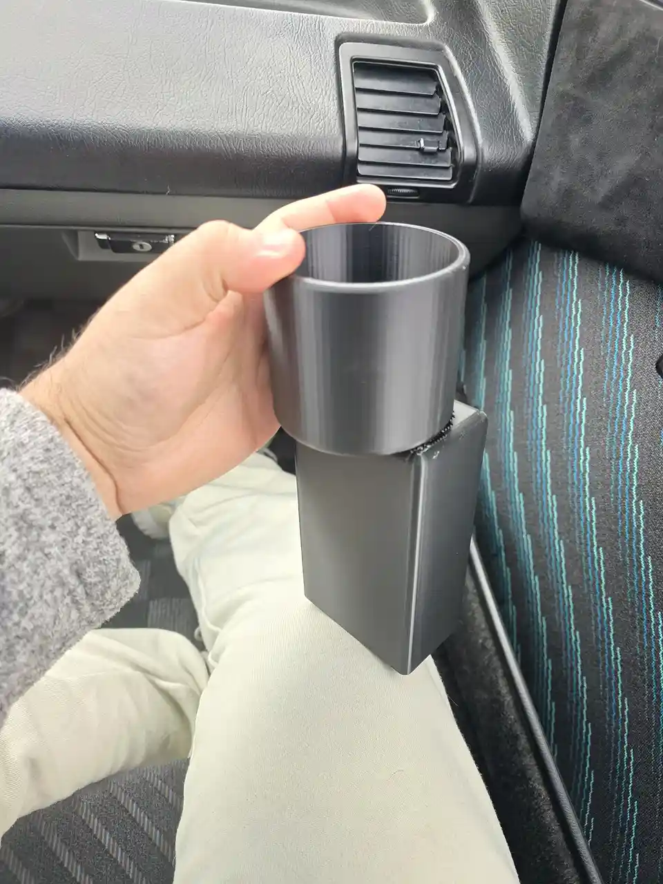 A person's hand holds a black 3d-printed cup holder attached to the car's center console, which is resting on their knee…