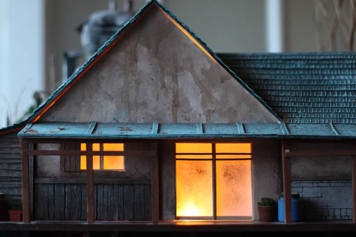 A detailed miniature model of a rustic house with glowing orange lights visible through its windows and along the roofline…