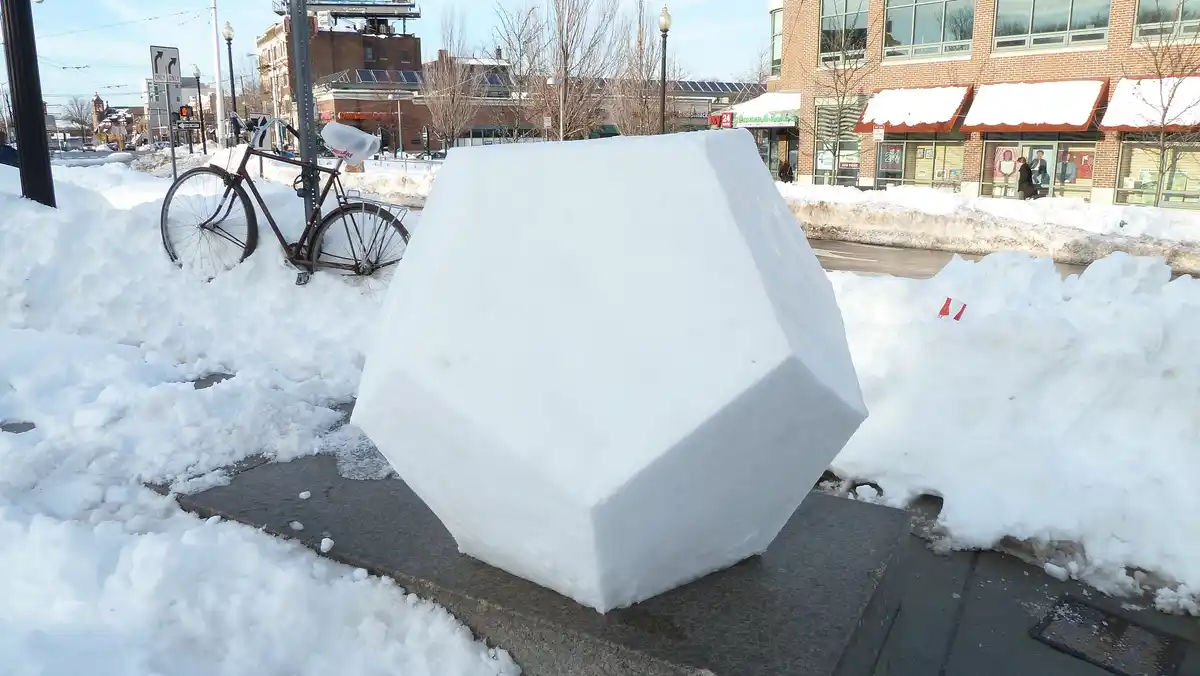 A large, geometrically shaped snow sculpture sits on a stone pedestal in a snowy urban sidewalk, with a bicycle partially…