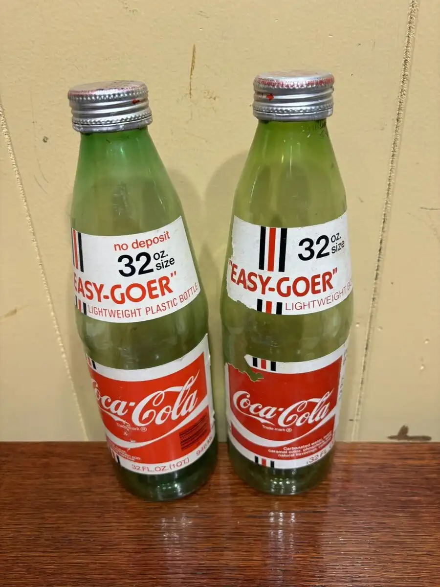 Two vintage green plastic Coca-Cola bottles, labeled 'EASY-GOER' and '32 oz. size', sit side-by-side on a wooden surface…
