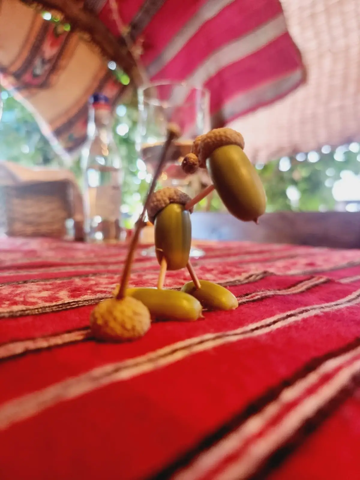 Two acorns with twig limbs and acorn caps are arranged on a red patterned tablecloth to appear as if one is leaning on the…