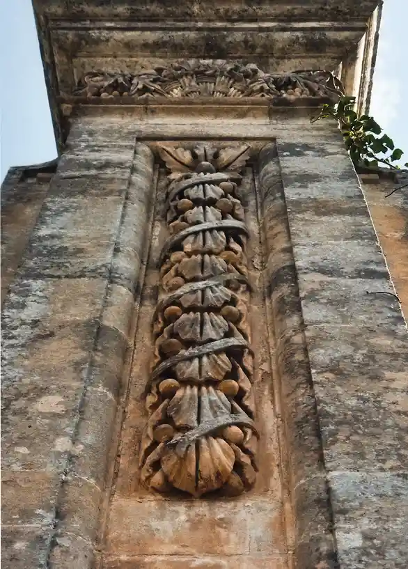 This image shows a close-up of a weathered stone architectural detail on an old building, featuring a vertical carved motif…