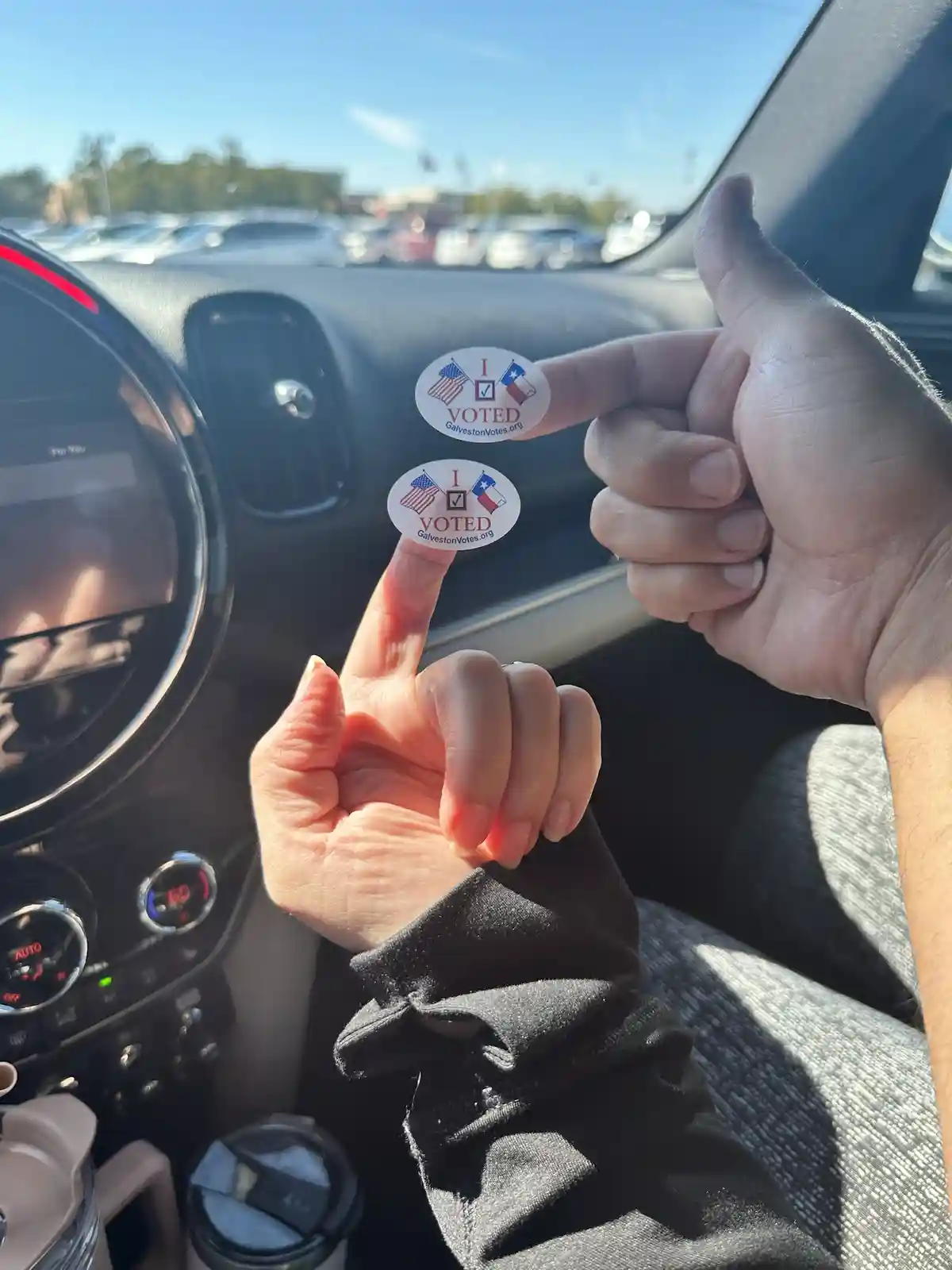 Inside a car, two people hold up their thumbs and index fingers, each displaying an 'I VOTED' sticker from…
