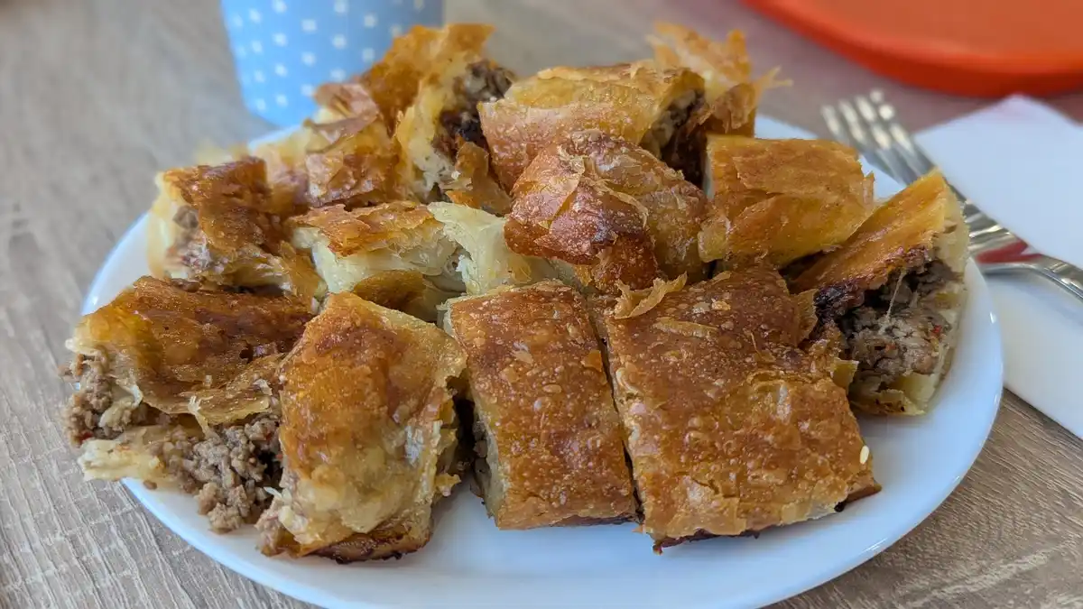 A close-up photo shows a white plate piled high with golden-brown, flaky, crispy meat-filled pastries, likely burek or a…