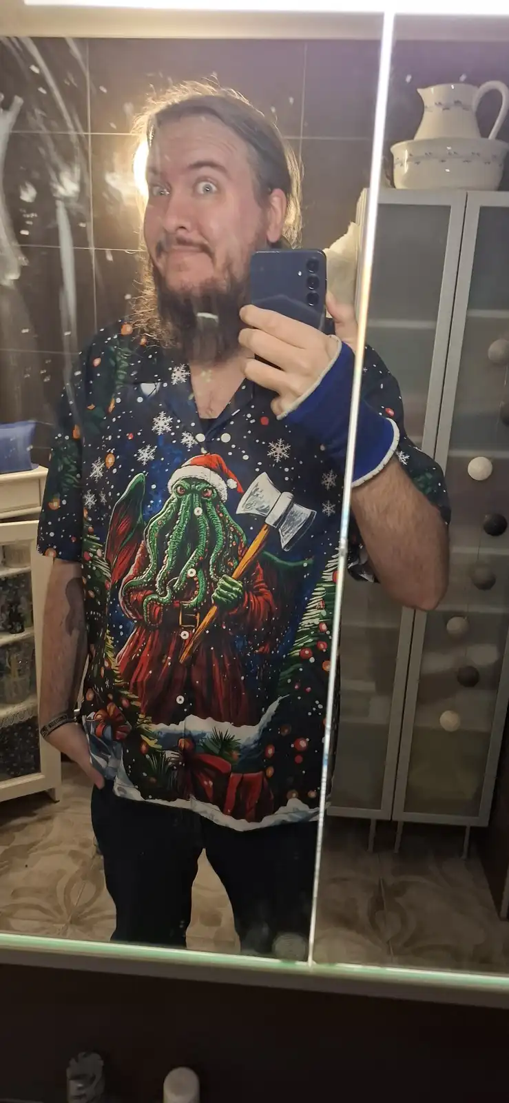 A man with a beard takes a mirror selfie in a bathroom, wearing a dark blue holiday-themed shirt featuring a green Cthulhu…