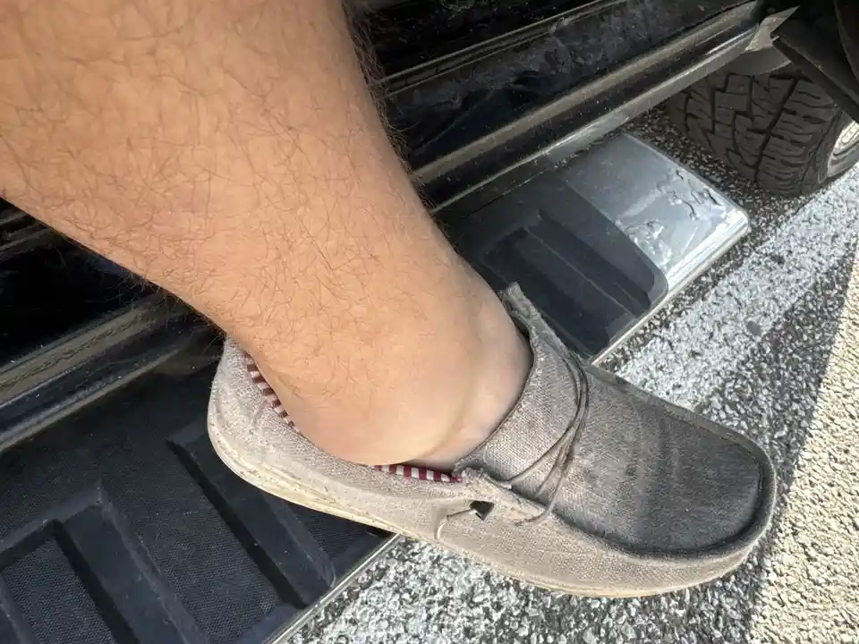 A close-up photo shows a person's hairy leg and foot wearing a worn gray slip-on shoe, resting on the black rubber floor mat…