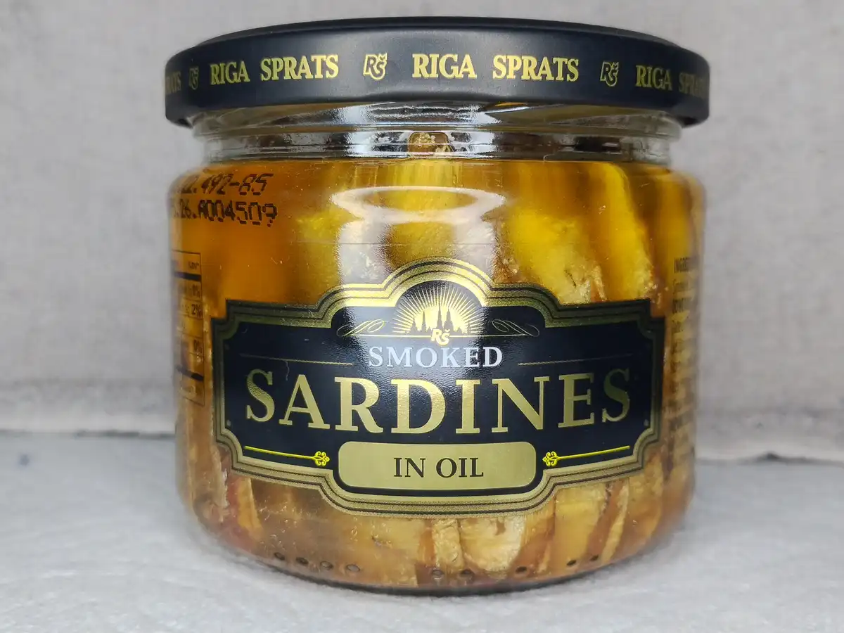 A close-up photo of a glass jar of Riga Sprats smoked sardines in oil, with a black and gold label and lid. The text on the…