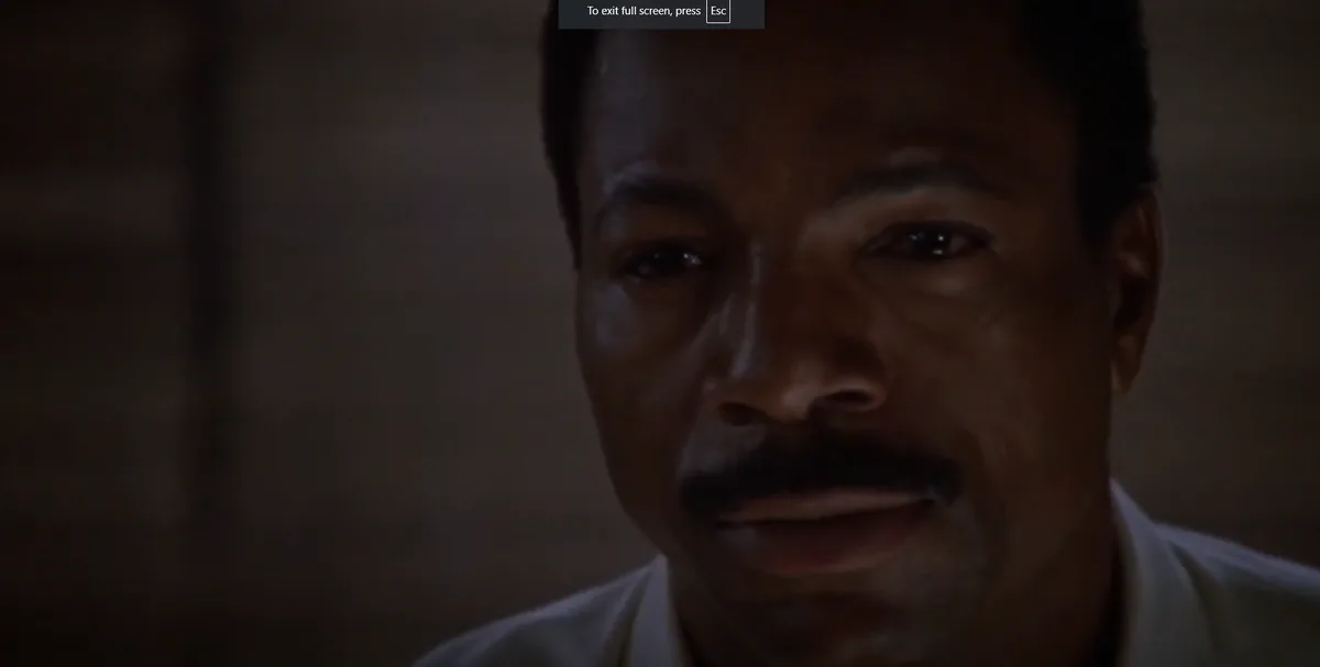 A close-up shot of a man with a mustache and a serious, intense expression, looking directly at the camera in a dimly lit…