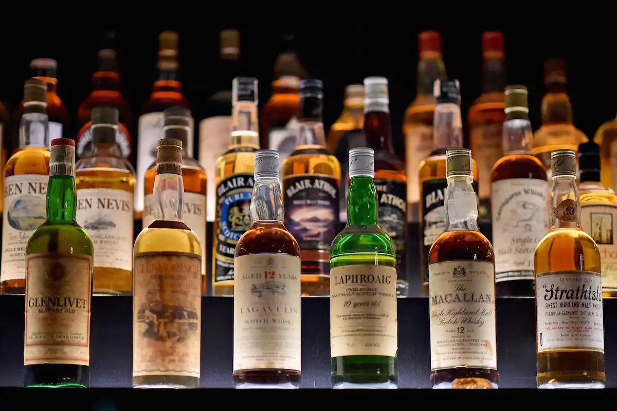 A row of various Scotch whisky bottles, including Glenlivet, Glenmorangie, Laphroaig, and The Macallan, are displayed on a…