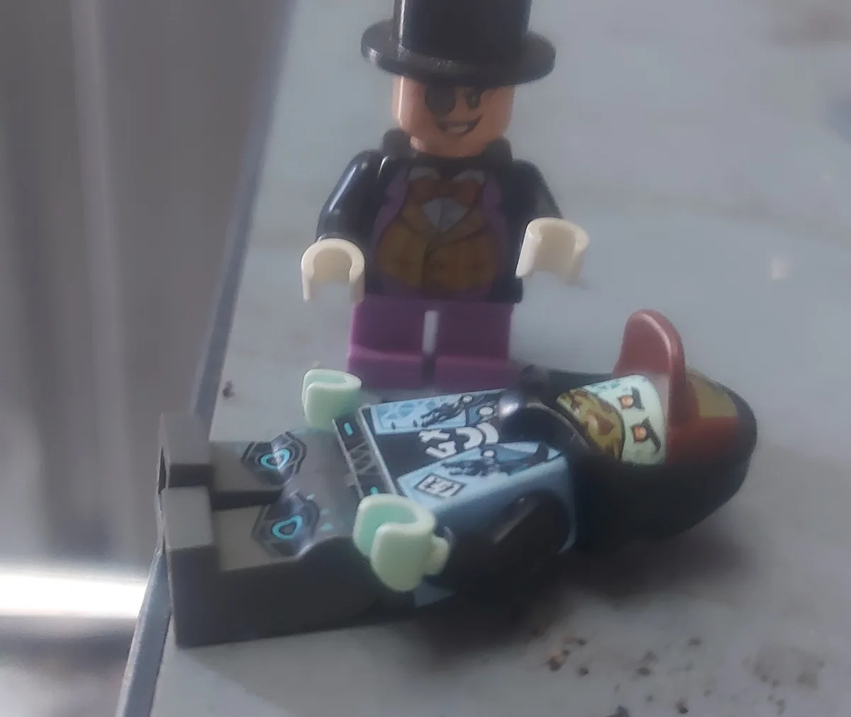 A lego figure dressed as the joker, wearing a black top hat and purple suit, stands over a lego batman figure lying on his…