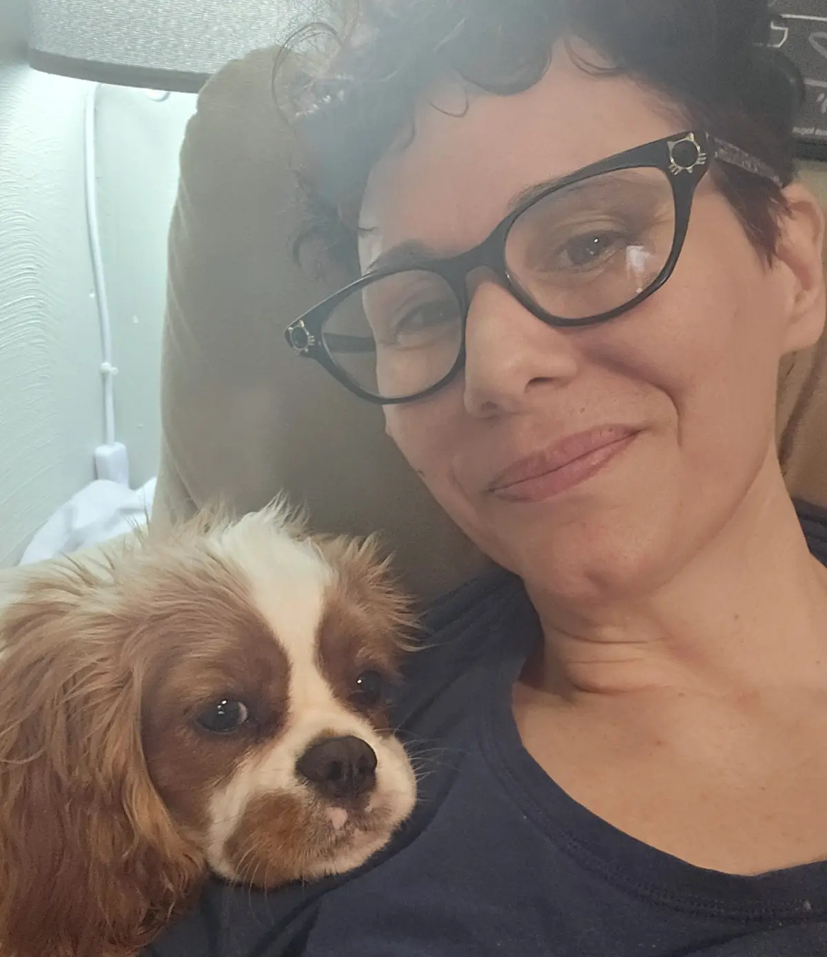 A woman with short dark hair and black-rimmed glasses smiles gently while holding a small brown and white Cavalier King…
