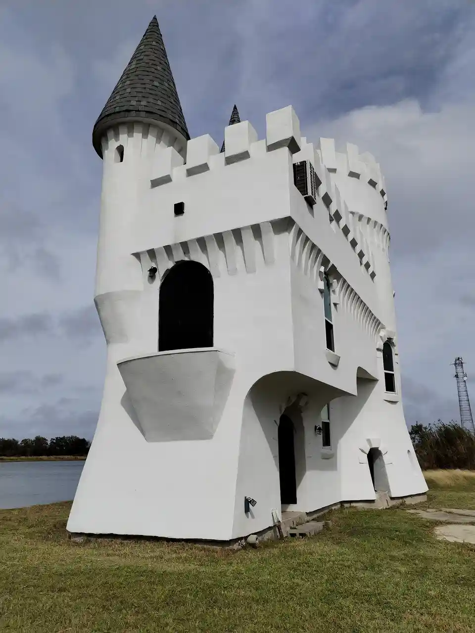 A white, castle-like structure with a conical roof and crenellated walls stands on grass near water under a cloudy sky. The…